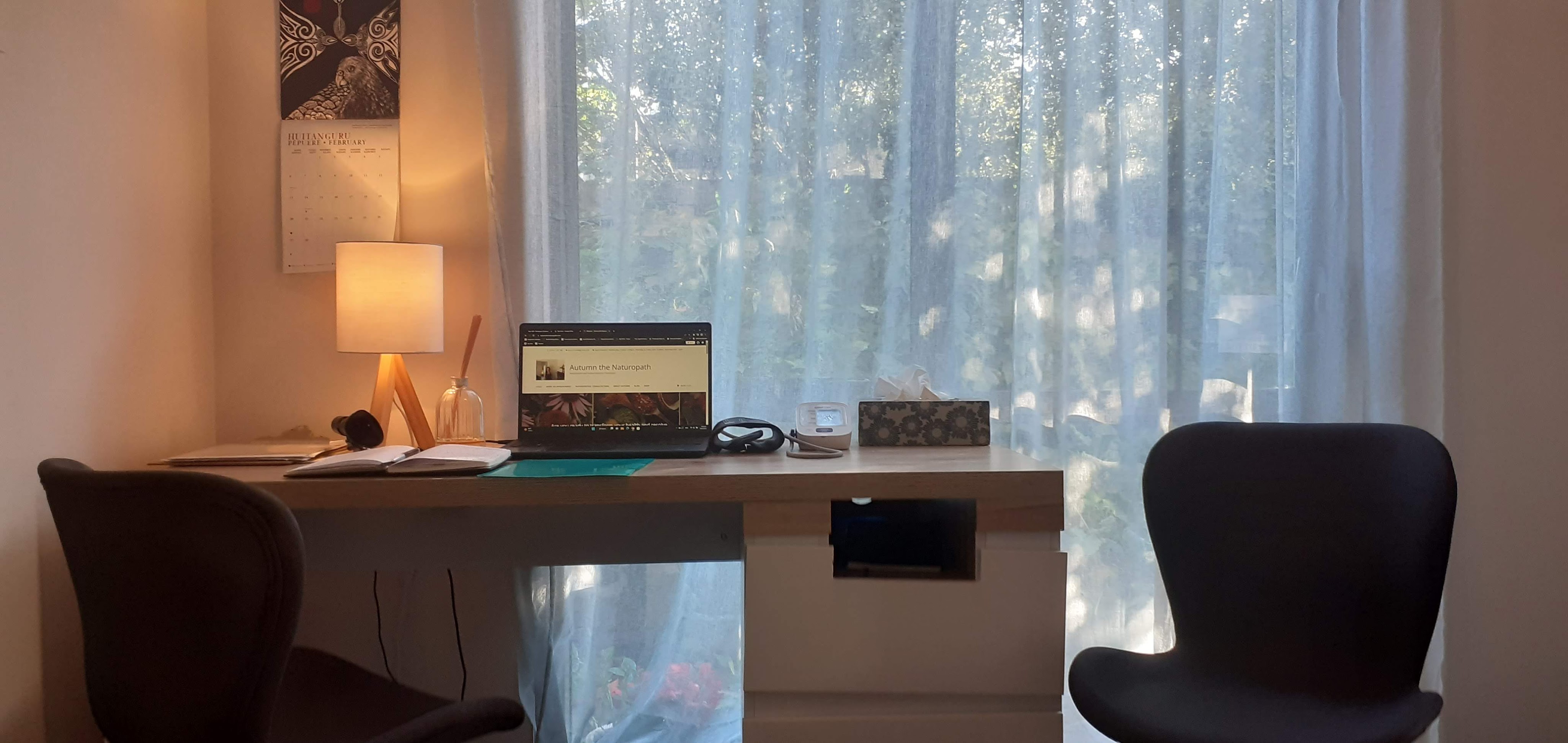 A cozy home office setup with a wooden desk, a laptop displaying a website, a lamp, and two chairs, illuminated by natural light from a nearby window with sheer curtains.