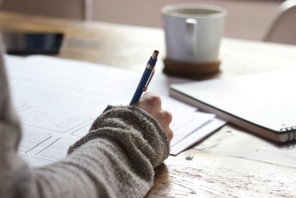 A person holding a pen writing on paper at a table with a cup on the table