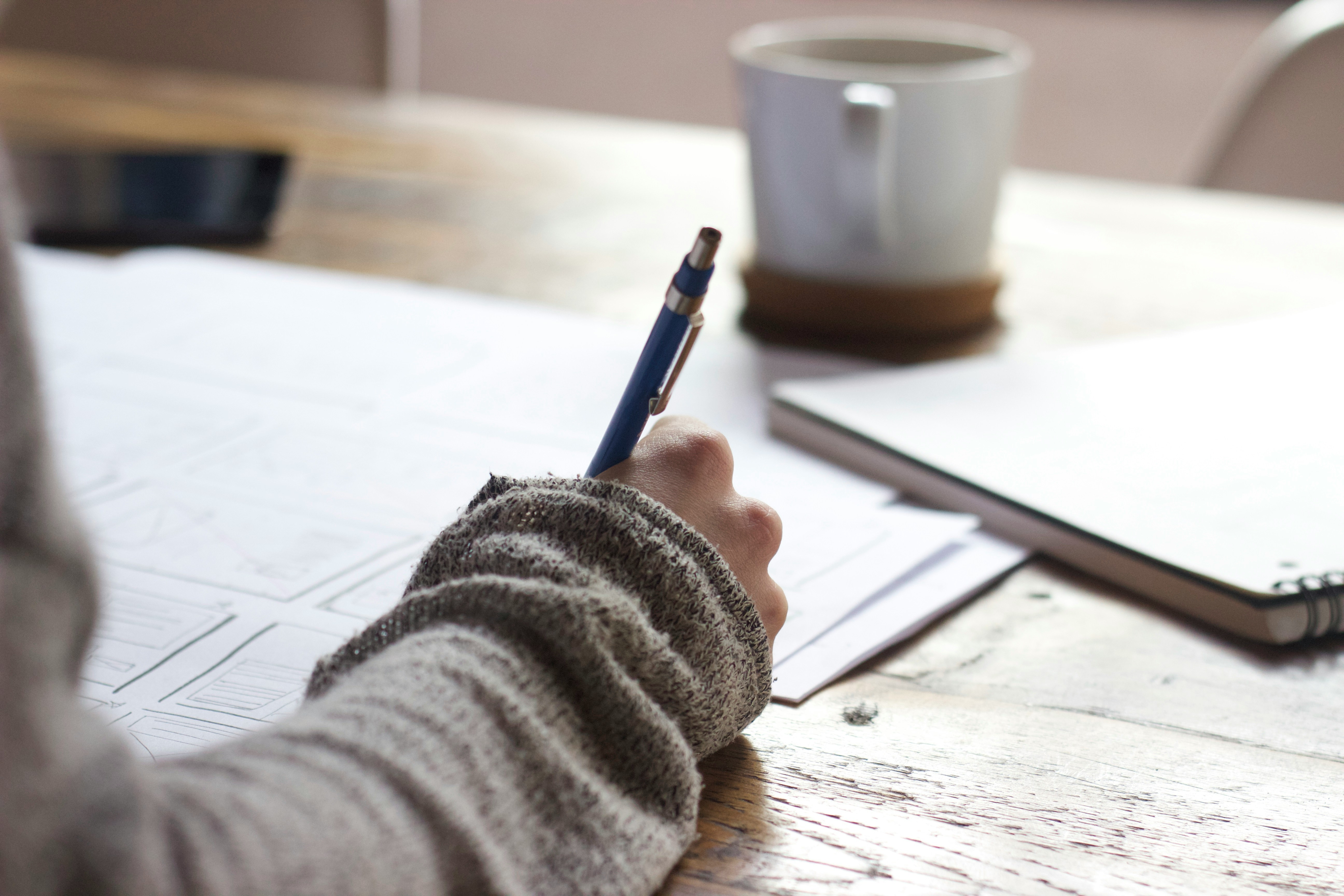 A person writing notes with a blue pen on paperwork at a wooden table, with a mug in the background.