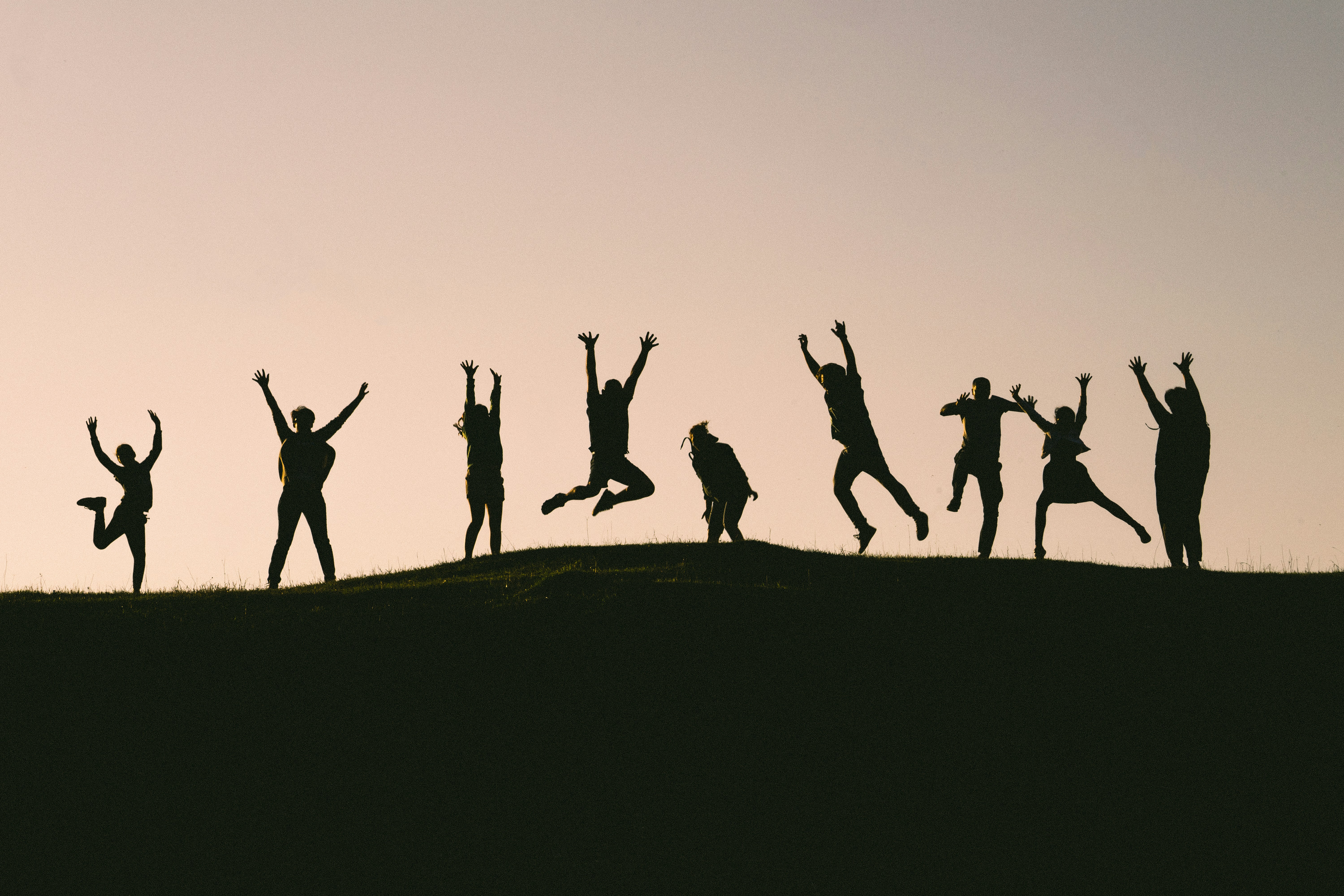 Silhouettes of a group of people joyfully jumping and posing on a hill at sunset.