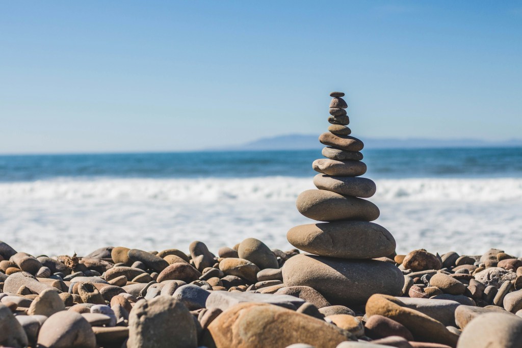 A stack of smooth stones balanced on a rocky beach, with the ocean waves gently rolling in the background under a clear blue sky.