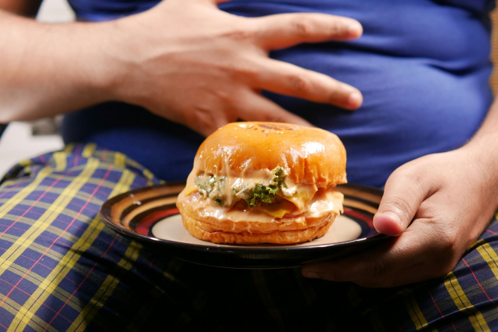 A close-up of a person's hand holding a burger on a plate, with their other hand resting on their stomach, indicating a potential discomfort or fullness.