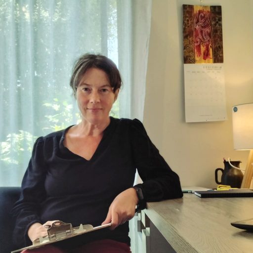 A woman sitting at a desk, holding a clipboard, with a bright window in the background and a calendar hanging on the wall.