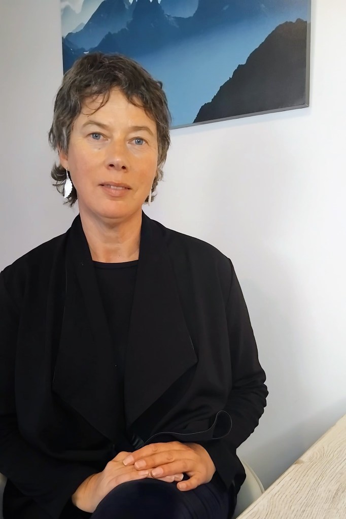 A woman with short, curly hair and earrings, sitting at a table and wearing a black outfit, with a mountain landscape painting in the background.