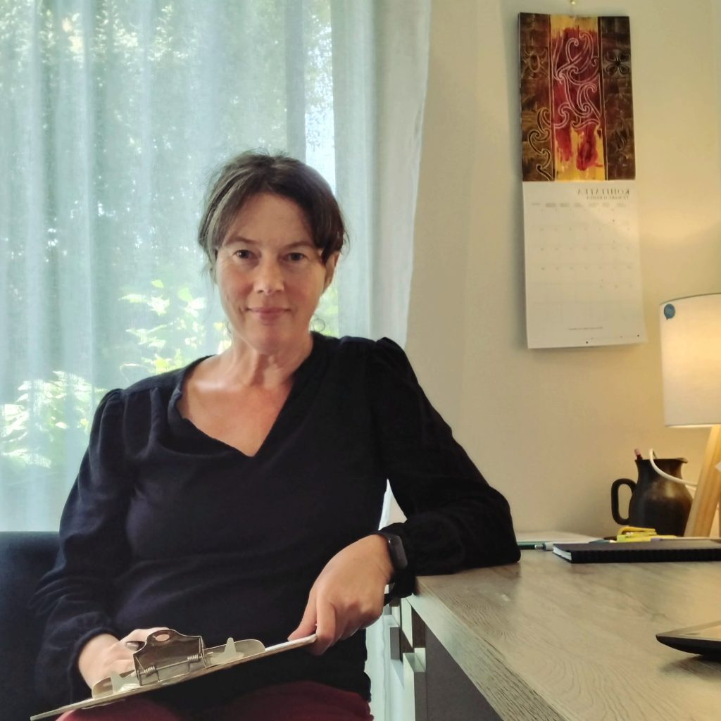 A woman sitting at a desk holding a clipboard. She has shoulder-length hair and is wearing a black top. The background features a window with light-colored curtains and a calendar on the wall.